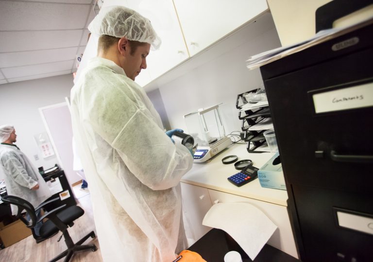 Young male lab tech in white disposable hat and coat pouring powder into stainless steel scoop