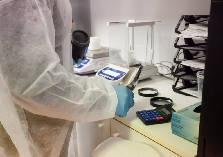 Lab tech in white disposable coat with blue latex gloves holding stainless steel scoop